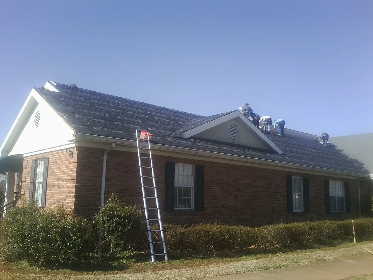 Crew tearing shingles off a brick ranch roof with a ladder set — Emergency Roof Repair work by our crew in Roxboro